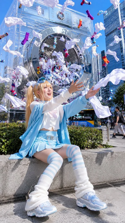 A seated person in a blue outfit raises an arm beneath floating objects and a large circular structure.